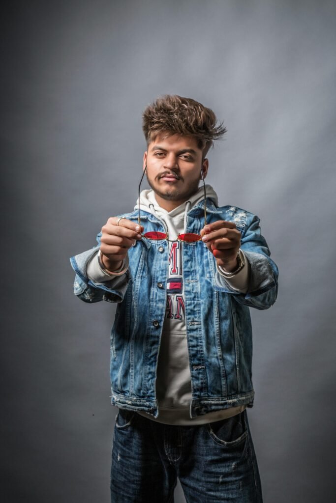 Young man in a denim jacket posing with sunglasses in a studio setting.